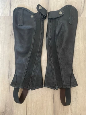 Svarta läderchaps från Equipage - Säljer ett par svarta chaps XS  i äkta läder från Equipage. De har dragkedja längs sidan med stretch och elastiska band under foten. Ger bra grepp och stabilitet utan att vara obekväma. Snygg och stilren design med tryckknapp upptill. Lädret putsats innan de skickas eller hämtas i Sjöbo. De är använda men bra skötta. Säljer då de är för små.. Nypris runt 600-800