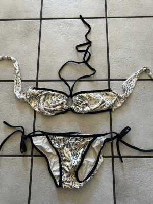 Leopardmönstrad bikini med knyt - Snygg bikini med leopardmönster i beige, svart och vitt. Överdelen har knyt i nacken och en ringdetalj framtill, medan underdelen har knyt i sidorna. Svarta kanter ger en cool kontrast. Perfekt för stranden eller poolen!