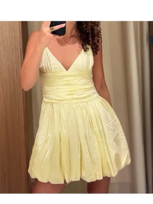 Ljusgul kort klänning med v-ringning - Superfin ljusgul kort klänning med v-ringning och omlottkänsla upptill. Klänningen har bred midja och voluminös kjol i glansigt material som ger en festlig look. Perfekt för sommarens alla tillfällen!