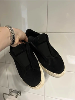 Svarta slip-on sneakers från Axel Arigato - Svarta slip-on sneakers från Axel Arigato med ovandel i mocka och elastiska paneler för enkel påtagning. Skorna har en vit, chunky gummisula och minimalistisk design utan snörning. Perfekta för en clean och stilren look.