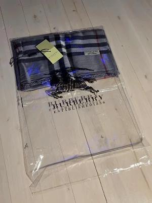 Grå-rutig halsduk från Burberry - Säljer en klassisk grå halsduk från Burberry med det ikoniska rutmönstret i svart, vitt och röda detaljer. Halsduken är tunn och har fransar i kanterna. Perfekt accessoar för att lyfta din outfit.