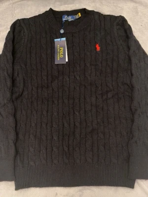 Svart kabelstickad tröja Polo Ralph Lauren - Kabelstickad svart tröja från Polo Ralph Lauren med rund halsringning och den klassiska röda logotypen broderad på bröstet. Tröjan har ribbade muddar vid ärmslut och nederkant. Perfekt för dig som gillar stilrena och tidlösa plagg.