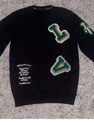 Svart sweatshirt louis Vuitton (LV) - Cool svart sweatshirt med stora gröna och vita bokstäver 'L' och 'V' på framsidan och ärmen. Broderad vit text på magen ger en unik detalj. Perfekt för dig som gillar streetwear och vill sticka ut med en trendig look.
