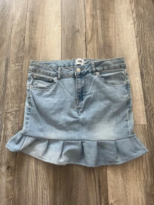 Ljusblå jeanskjol med volangkant - Snygg ljusblå jeanskjol med klassiska fickor fram och bak samt en trendig volangkant nedtill. Kjolen har bälteshällor och knappstängning framtill. Perfekt för dig som gillar en cool och avslappnad stil.