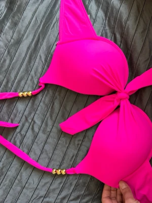 Rosa bikini med gulddetaljer - Snygg bikini i starkt rosa färg med knyt framtill och guldfärgade pärldetaljer på banden. Bikinin har vadderade kupor och trekantsmodell, perfekt för sommaren och strandhäng.Helt ny