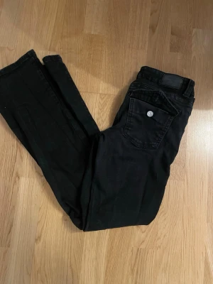 Svarta jeans med ficklock - Säljer ett par svarta jeans med utvida ben och snygg ficka med lock och silverfärgad knapp bak. Jeansen har klassisk femficksdesign och är tillverkade i denim med stretch för skön passform.
