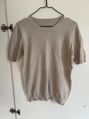 Beige kortärmad stickad tröja - En stilren beige stickad tröja med korta ärmar och rund halsringning. Tröjan har ribbade muddar vid ärmslut och nederkant, vilket ger en snygg passform. Perfekt för lager-på-lager och enkel att matcha med olika outfits.