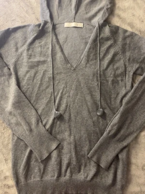 Grå hoodie med v-ringning - Mysig grå hoodie med v-ringad hals och långa ärmar. Tillverkad i mjukt, tunt stickat material och har en huva med dragsko och små bollar i ändarna. Perfekt för en avslappnad och chill stil.
