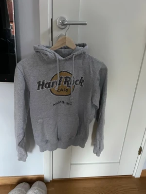 Grå Hard Rock Cafe hoodie - Grå hoodie från Hard Rock Cafe med tryck från Hamburg på bröstet. Klassisk känguruficka framtill, dragsko i huvan och ribbade muddar. Perfekt för dig som gillar streetwear och vill ha en chill look. Materialet är mjukt och bekvämt, troligen bomullsblandning.