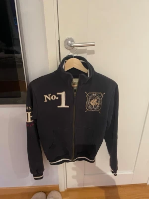 Mörkblå hoodie från Sports Wear Equestrian - Mörkblå hoodie med dragkedja och vita broderade detaljer som 'No.1' och klubbmärke fram samt 'S.W.E.' på ryggen. Ribbstickade muddar och krage, samt fickor fram. Perfekt för en sportig och avslappnad stil.