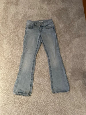 Ljusblå bootcut jeans - Säljer ett par ljusblå bootcut jeans med klassisk femficksdesign. Jeansen har en något utsvängd passform nertill och är tillverkade i denim. Perfekta för en avslappnad och trendig look. Det är ett litet hål på ett av benen men det är ingenting som syns.