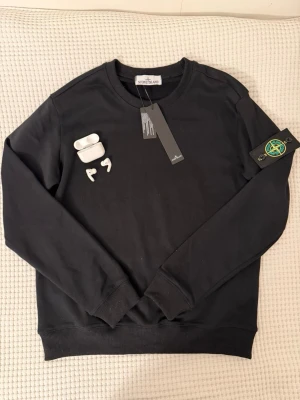 Svart sweatshirt från Stone Island - Svart sweatshirt från Stone Island med klassisk rund hals och ribbade muddar. Ikonisk logotyp-patch på vänster ärm. Tröjan är i mjukt material och har en stilren, enkel design som passar perfekt till streetwear-stilen.passar M och S