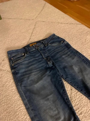 Jack & Jones Slim Glenn jeans blå - Snygga blå jeans från Jack & Jones, modell Slim Glenn. De har en slim passform och klassisk femficksdesign med slitningar och tvättade detaljer. Jeansen är tillverkade i jeansmaterial och har knapp- och dragkedjestängning.