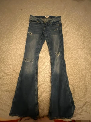 Blåa bootcut jeans från Gina Tricot - Säljer ett par blåa bootcut jeans från Gina Tricot med slitna detaljer och hål på benen. Jeansen har låg midja, klassisk femficksmodell och är tillverkade i denim. Perfekta för dig som gillar en retro och avslappnad stil.