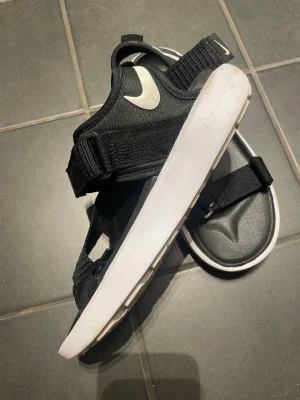 Svarta Nike sandaler med vita sulor - Snygga svarta sandaler från Nike med justerbara remmar och vit sula. Sandalerna har en sportig look med tydlig Nike-logga på remmarna och sidan. Tillverkade i textil och syntetmaterial för en bekväm känsla hela dagen.