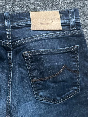 Mörkblå jeans från jacob cohen - Säljer ett par klassiska mörkblå jeans från jacob cohen med raka ben och femficksmodell. Jeansen har kontrastsömmar och en läderpatch med logga bak i midjan. Perfekta till en avslappnad stil och tillverkade i slitstark denim.