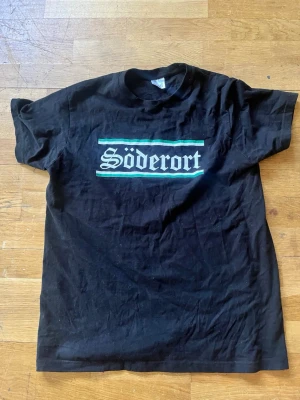 Svart t-shirt med Söderort-tryck - Svart t-shirt med trycket 'Söderort' i vit gotisk stil på bröstet, inramat av gröna och vita linjer. Klassisk rund hals och korta ärmar. Perfekt för dig som vill representera Söderort med stil.