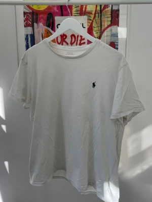 Vit t-shirt från Polo Ralph Lauren - Klassisk vit t-shirt från Polo Ralph Lauren med rund halsringning och den ikoniska mörkblå logobroderingen på bröstet. Tillverkad i mjuk bomull och har en normal passform. Perfekt basplagg för alla tillfällen. Storlek L men passar även övre M.