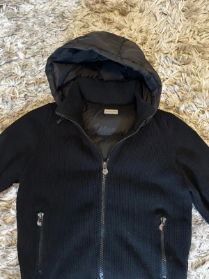 Svart cardiganjacka från Moncler - Svart stickad cardiganjacka från Moncler med quiltad huva och innerfoder. Jackan har dragkedja framtill, två fickor med dragkedja och Moncler-logga på ärmen. Perfekt för kyliga dagar och snygg till streetwear.