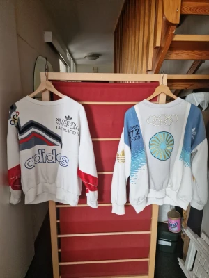 Vintage Adidas unik OS-tröjor med retrotryck - Två vita vintage Adidas-tröjor med färgglada retrotryck från OS, bland annat Lake Placid 1980 och München 1972. Tröjorna har rund hals, långa ärmar och detaljer i blått, rött, svart och gult. Perfekt för dig som gillar sportig och unik stil.