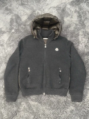 Äkta Moncler cardigan - Mörkgrå stickad cardiganjacka från Moncler med olivgrön quiltad dunhuva och rygg. Storlek L men passar runt 170cm. Jackan är 100% äkta då det inte ens finns kopior på denna exakta modell. Jackan har två dragkedjefickor fram, ribbade muddar och Moncler-logga på bröstet. 