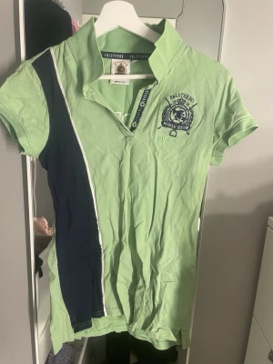 Grön Falsterbo pikétopp med tryck - Grön pikétopp från Falsterbo Horse Show med mörkblå och vit diagonal detalj framtill. Broderad logga på bröstet och stort 'CREW'-tryck på ryggen. Klassisk krage och korta ärmar. Perfekt för dig som gillar hästtema och sportig stil.