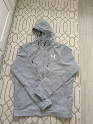 Ljusgrå vindjacka från Under Armour - Snygg ljusgrå vindjacka från Under Armour med huva och dragkedja framtill. Jackan har två sidofickor, elastiska muddar och meshfoder för extra komfort. Perfekt för blåsiga dagar och sportiga outfits.