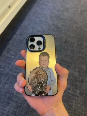 Mobilskal med guld och rolig bild - Unikt mobilskal med spegelblank guldfinish och tryck av ett barn som håller ett stort mästarbälte. Skalets yta är reflekterande och ger en cool look till din mobil. Passar perfekt för dig som vill sticka ut med ett personligt och humoristiskt skal.