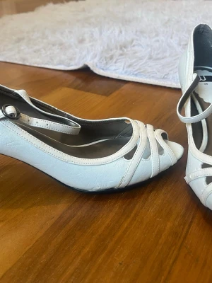 Vita 2000s klackar med rem - Snygga vita 2000s pumps med peep toe och smal rem. Skorna har dekorativa remmar framtill och en klassisk klack. Perfekta för dig som gillar stilrena och feminina skor.