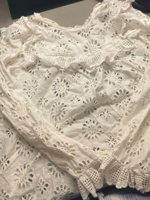 Vit broderad blus från Zara Woman - Söt vit blus från Zara Woman med broderade blommönster och hålbroderier. Blusen har långa ärmar med elastiska muddar och volangdetaljer vid axlar och ärmslut. Tillverkad i ett lätt och luftigt material, perfekt för en trendig och romantisk look.
