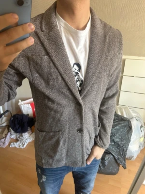 John henric kavaj med enkelknäppning - Stilren gråmelerad kavaj med klassisk passform och enkelknäppning. Kavajen har två framfickor och mjukt tyg med diskret struktur. Perfekt för en snygg och avslappnad look.