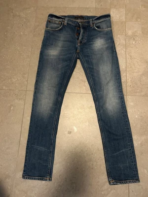 Nudie jeans tilted tor - Snygga blå slim fit jeans från Nudie med klassisk femficksdesign och orangea sömmar. Jeansen har en lätt tvättad look med slitningar på låren och bakficka med Nudie-logga. Tillverkade i bomull med knappgylf. W31/l 32 model tilted tor