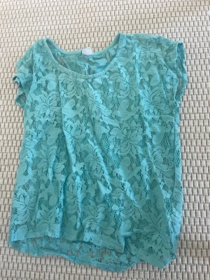 Turkos spetsblus från Gina Tricot - Snygg turkos blus från Gina Tricot i genombruten spets med blommigt mönster. Blusen har rund halsringning och korta ärmar, perfekt för en fräsch och somrig look. Luftig och lätt att matcha med jeans eller kjol.