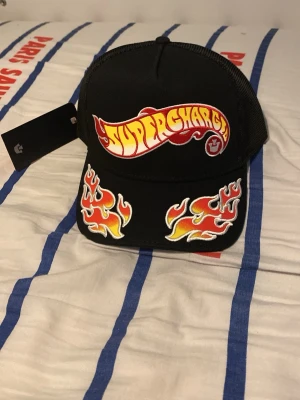 Svart keps med Supercharge och flames - Helt ny, har inte haft på mig någonting. Pris kan diskuteras. Cool svart keps med mesh baktill och broderad Supercharge-logga i rött och gult framtill. Skärmen har två stora flame-motiv i orange, gult och vitt som ger kepsen en riktigt edgy look. Perfekt för dig som gillar streetstyle och unika accessoarer.