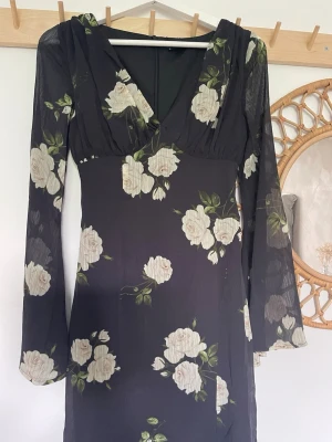 Svart långklänning med blommor - Svart långklänning med vita blommor och gröna blad. Klänningen har v-ringning, långärmade ärmar och figurnära passform. Tunt, lätt material.