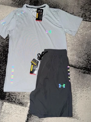 Under Armour  - Snyggt träningsset från Under Armour med en ljusgrå t-shirt och svarta shorts. T-shirten har färgglada loggor och detaljer, samt korta ärmar och V-ringning. Shortsen har dragsko i midjan och matchande logga. Perfekt för gym eller löpning.