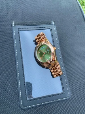 Rosé guld Nibosi klocka med oliv grön urtavla - Snygg klocka från Nibosi med rosé guldig länk och oliv grön urtavla. Klockan har tydliga index, dag- och datumvisning samt en robust metallarmband. Perfekt accessoar för dig som vill sticka ut med stilren design och cool färgkombo. Man får även med ett litet verktyg för att kunna justera klockan till sin önskade storlek. Rostfritt stål! 