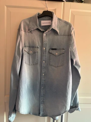 Ljusblå jeansskjorta från Calvin Klein - Snygg ljusblå jeansskjorta från Calvin Klein med klassisk krage, två bröstfickor med knappar och logotypdetalj. Skjortan har långa ärmar och knäppning framtill. Perfekt för en avslappnad och trendig look.