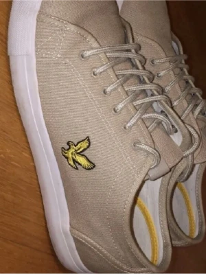 Beige sneakers från Lyle & Scott - Snygga beige sneakers från Lyle & Scott med klassisk låg profil och vit sula. Skorna är i canvasmaterial och har det ikoniska gula fågelbroderiet på sidan. Perfekta för en clean och stilren look. Skosnören i matchande beige färg.