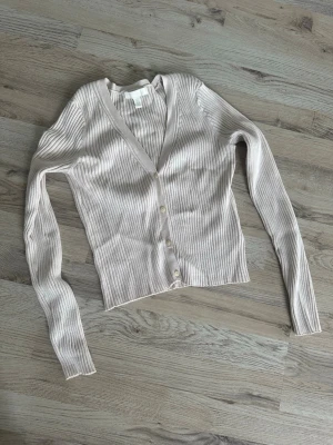 Beige ribbad kofta från H&M - Säljer en beige ribbad kofta från H&M med v-ringning och långa ärmar. Koftan har knäppning framtill med fyra små knappar och är tillverkad i ett mjukt, stretchigt material. Perfekt att slänga över en topp för en avslappnad look.