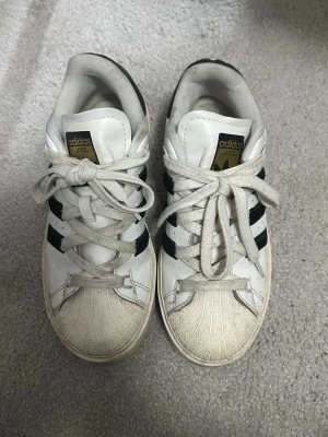 Adidas Superstar vita sneakers - Klassiska Adidas Superstar sneakers i vitt läder med svarta ränder och guldiga detaljer på tungan och sidan. Skorna har en ikonisk snäcktå-design och tjock vit sula. Perfekt för dig som gillar retrostil och streetwear.
