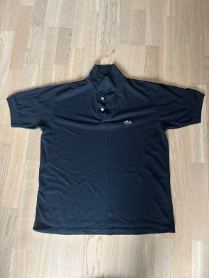 Svart pikétröja från Lacoste - Klassisk svart pikétröja från Lacoste med korta ärmar och krage. Tröjan har två knappar framtill och den ikoniska krokodilloggan broderad på bröstet. Perfekt till en avslappnad men ändå stilren look.