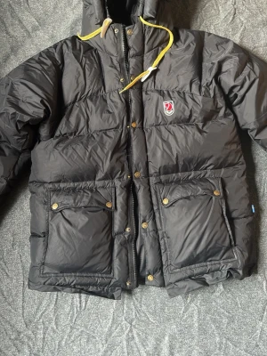 Svart dunjacka från Fjällräven - Svart dunjacka från Fjällräven med huva och gula dragsnören. Jackan har två stora fickor framtill med lock och knappar, samt dragkedja och tryckknappar. Klassisk Fjällräven-logga på bröstet. Perfekt för kalla vinterdagar, skidresor och kallt väder. Pris kan diskuteras!