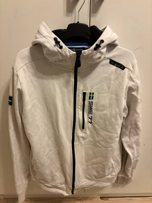 Vit sail racing SWE 77 hoodie med huva - Vit hoodie från Sail racing med dragkedja, huva med snörning och svart innerfoder. På bröstet finns svensk flagga och texten SWE 77, samt en svensk flagga på ärmen. Materialet är mjukt och bekvämt, perfekt för chill dagar. Reflektiv detalj på huvan.
