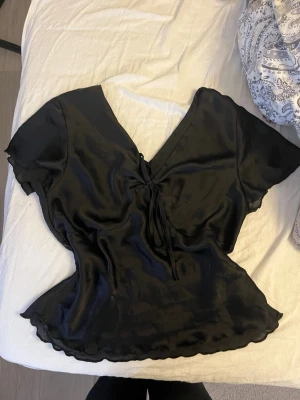 Svart omlottopp i satinlook - Snygg svart omlottopp med korta ärmar och v-ringning. Toppen har en knytning framtill och är gjord i ett glansigt satinliknande material som ger en lyxig känsla. Perfekt att styla med jeans eller kjol för en trendig look.