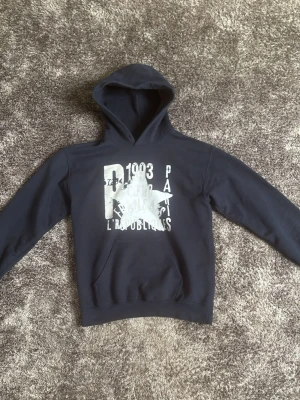 Mörkblå hoodie Zandro Studio - Mörkblå hoodie med stor huva och magficka. Framsidan har ett vitt tryck med text och grafiska detaljer, bland annat siffror och ordet 'Paris'. Tillverkad i mjukt sweatshirtmaterial, perfekt för en avslappnad stil.