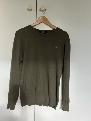 Olivgrön stickad tröja Polo Ralph Lauren - Snygg olivgrön stickad tröja från Polo Ralph Lauren med rund halsringning och diskret broderad logga i guld på bröstet. Tröjan har långa ärmar och ribbade muddar vid ärmslut och nederkant. Perfekt till ett avslappnat men stilrent uttryck. 15% Kashmir.