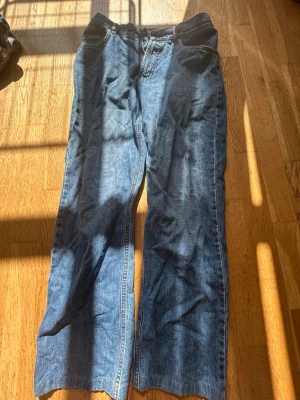 Blå raka jeans med svarta detaljer - Säljer ett par blå jeans med raka ben och svarta detaljer upptill vid midjan. Klassisk femficksmodell med normalhög midja och dragkedja. Jeansen har en avslappnad passform och är tillverkade i denim. Perfekta för en chill och stilren look.