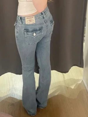 Ljusblå bootcut jeans från True Religion - Säljer ett par ljusblå bootcut jeans från True Religion med ikoniska sömmar och ficklock bak. Jeansen har en klassisk femficksmodell och silverfärgade detaljer på fickorna. Perfekta för dig som gillar en avslappnad men ändå trendig look. 