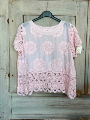 Ljusrosa blus med spetsdetaljer - Supersöt ljusrosa blus med korta ärmar och stora virkade spetsblommor över hela plagget. Blusen har rund halsringning och en lös passform, perfekt för en romantisk look. Materialet känns luftigt och lätt.